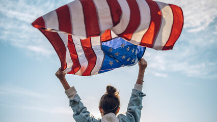 Portrait of a young beautiful woman in glasses and jeans with a waving American flag in her hands,...