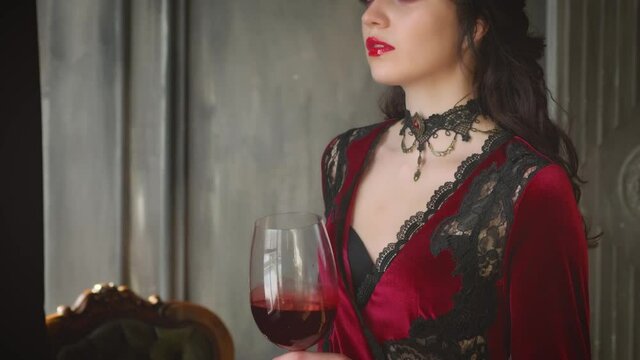 Closeup portrait gothic beautiful mystical vampire woman. Brunette Queen holding glass blood wine in hands. Luxury vintage red dress. Black necklace. Art mystic makeup. Dark walls old castle, armchair