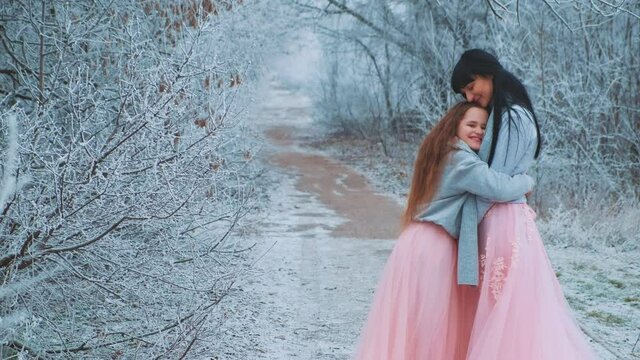 Joyful cute pretty little girl daughter hugs young beautiful woman mom. Concept happy childhood. Same clothes blue warm sweater, pink long luxury skirt dress. Loose redhaired black hair. Snowy forest 
