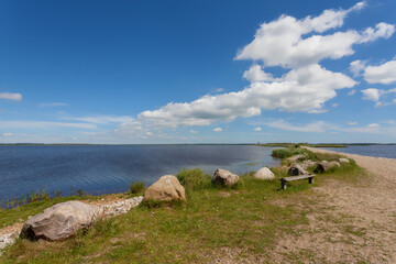 big stones and a simple wooden bench at the lake Filsø (Denmark) on a sunny summer day with blue water and sky with scenic white clouds