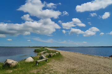 a simple wooden bench without backrest and some boulders stand at a dam in the lake Filsø on a sunny summer day with scenic white clouds on blue sky