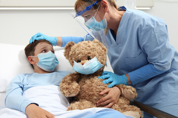 nurse takes care of the patient child in hospital bed playing with teddy bear, wearing protective...