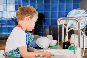 a boy in an apron washes dishes in the kitchen at home.