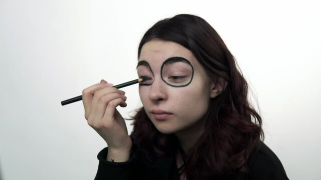Woman using pencil to draw black circles to make santa muerte makeup. The girl is sitting in the studio and applies black paint around eyes. Theme of santa muerte carnival and sugar skull makeup.