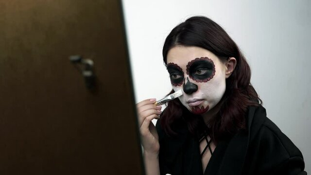 Woman applies white bodyart paint at her face for santa muerte makeup. The girl in the studio uses brush to apply the paint over her face. Theme of death day and it's traditional sugar skull makeup.