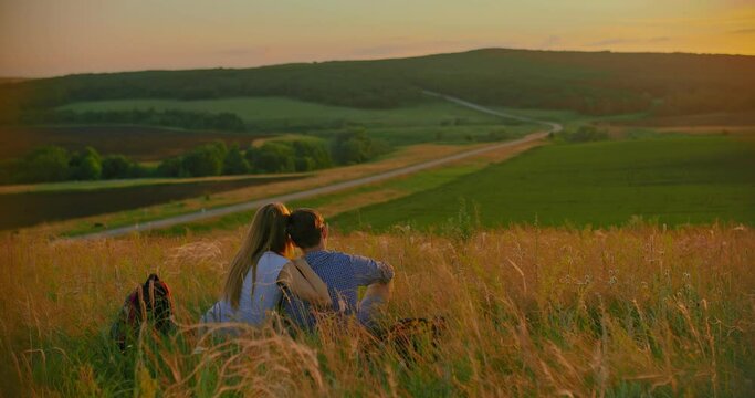 Pair of tourists in love sits on a hill and enjoys the sunset. Road goes into the distance, green fields. View from the back.