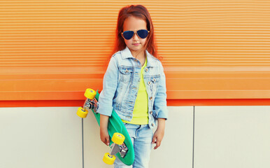 Stylish little girl child with skateboard on city street over orange wall background © rohappy