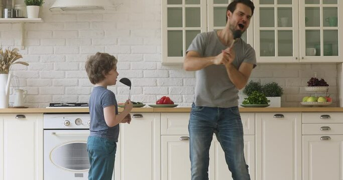 Funny artistic father and cute little kid son singing song in kitchen, happy family dad with child boy holding spoon ladle kitchenware microphones enjoy karaoke dancing having fun in kitchen together