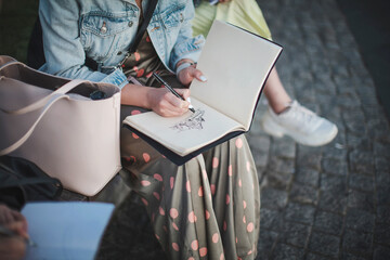 Young beautiful girl draws in a park in a group. Outline in a sketchbook.