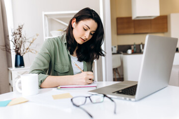 Online education. A young attractive caucasian student girl writes information in her notebook during online learning at home