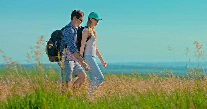 Pair of lovers is walking across the field with backpacks and holding hands. Hike, clear blue sky and grass swaying in the wind.