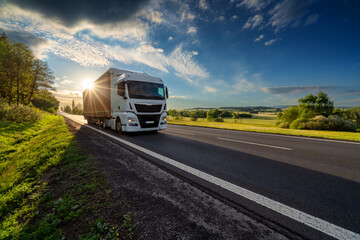 Fototapeta na wymiar White truck driving on the asphalt road in rural landscape at sunset