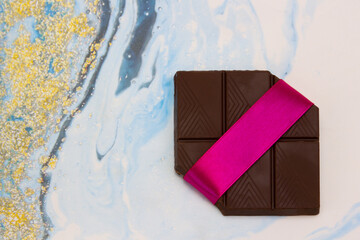 A piece of chocolate tied with a gift ribbon on a multicolored light background, top view.