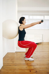 Woman pressing fitness ball against the wall while lifting dumbbells