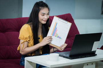 Naklejka premium A young Asian female university student presents the chart on her project study online via the laptop at home during the COVID-19 pandemic to practice social distancing. Stock photo.