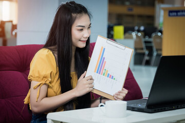 A young Asian female university student presents the chart on her project study online via the laptop at home during the COVID-19 pandemic to practice social distancing. Stock photo.