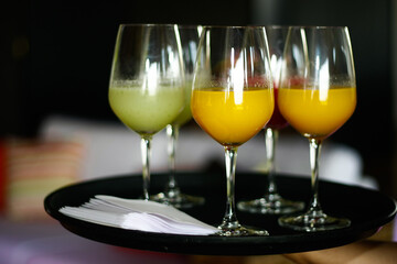 Taças de vidro contendo sucos de laranja e abacaxi com hortelã, dispostas sobre uma bandeja preta com guardanapos de pano brancos.