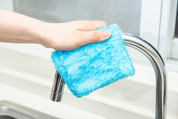 A hand holding a blue cleaning cloth is cleaning the faucet in the sink.