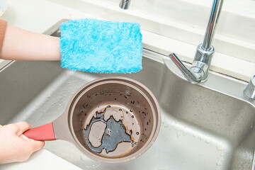 A hand in front of the sink with a blue cleaning cloth, cleaning a greasy soup pot. 