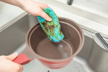 A hand in front of the sink with a blue cleaning cloth, cleaning a greasy soup pot. 