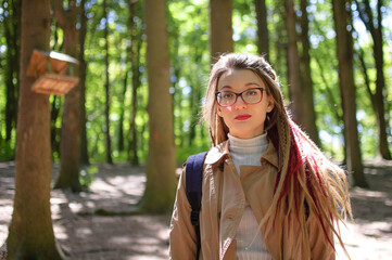 Young woman wearing glasses, casual nude trench, and dreadlocks is standing at the urban park on trees background during sunny spring day outdoors