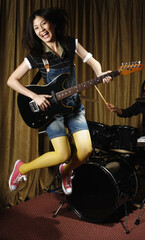 Young woman jumping while playing guitar, man playing drums in the background