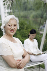 Senior woman folding her arms while smiling at the camera, woman reading book in the background