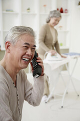 Senior man talking on the phone, senior woman ironing clothes in the background