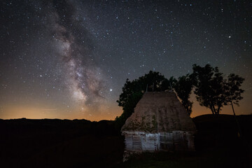 Old abandoned barn farm house with some trees behind it shot in the night against a starry sky with milky way galactic core seen above