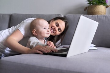 Young smiling mom with toddler baby look together at laptop