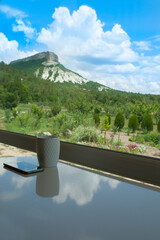 Cup of coffee and mobile phone on a table by the window overlooking the garden, mountain, forest and white clouds against a blue sky, sunny summer day. Copy space. Freelance, travel, remote work.