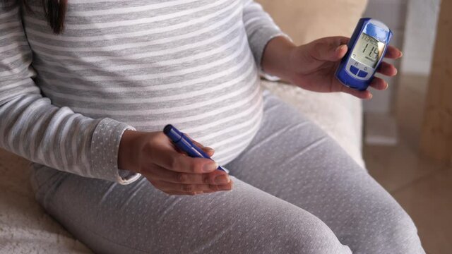 Portrait of a pregnant woman with a blood glucose meter in her hands at home on the bed. A pregnant woman with diabetes measures her blood glucose level at home while sitting on a bed.