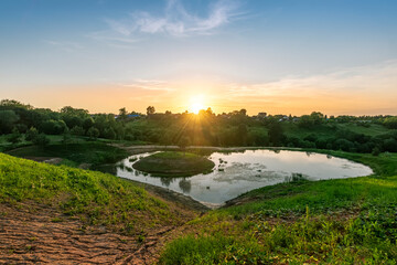 Scenic view at beautiful spring sunset with reflection on a shiny lake in shape of heart, grass, golden sun rays, calm water ,deep blue sky and glow on a background, spring evening landscape