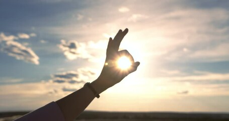 A silhouette of a close-up hand shows an ok gesture against a sunset beautiful sky with clouds and sun. Between the fingers that formed a circle, the sun is visible.