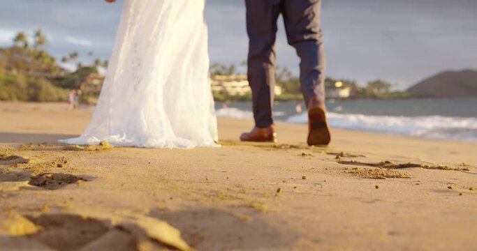 Just married Caucasian couple walk down white sandy hawaiian beach during golden hour. Close up of feet/legs. Shallow DOF