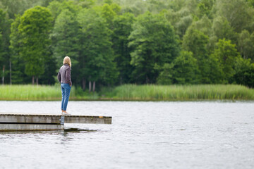 Fototapeta na wymiar One young adult woman standing alone on edge of footbridge and staring at lake and green trees in summer day. Thinking about life. Spending time alone in nature. Peaceful atmosphere. Back side view.
