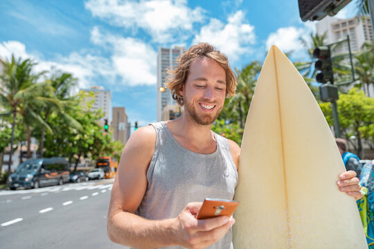 Mobile phone young man surfer going surfing walking in city with surfboard texting online on smartphone.