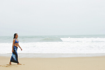 A lady in bikini top and wraparound skirt walking bare feet along the beach