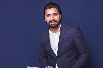 Indian businessman looking at camera, sitting with laptop over blue studio background