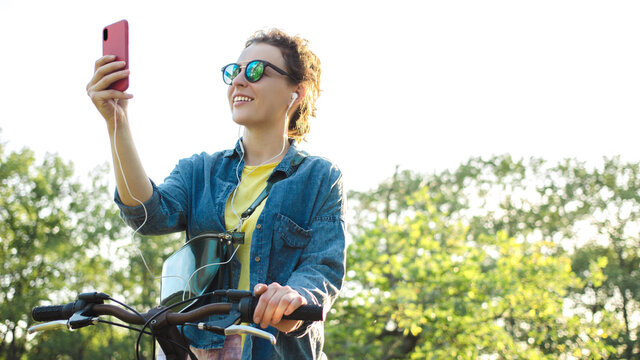 Smiling girl in sunglasses, headphones, holding smartphone,riding bicycle in park forest outdoor. Happy brunette woman sharing activity data on social media, using wireless internet connection 4g 5g