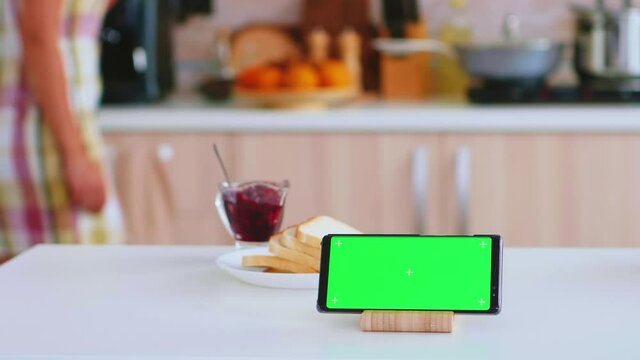 Smartphone with green mock up next to french pastery in kitchen and woman leaving cup of coffee on table top. Green screen chroma mock up isolated mockup background ready to be replaced with your text