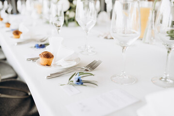 Decorated table with white tablecloths, cutlery, glasses, plates with napkins, next to a festive small bouquet of flowers with a blue blossoms. Themed minimalist table decor. Wedding or birthday party
