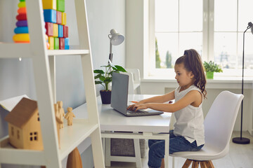 Online education of children. Serious little girl is typing laptop text while sitting at a table at home.