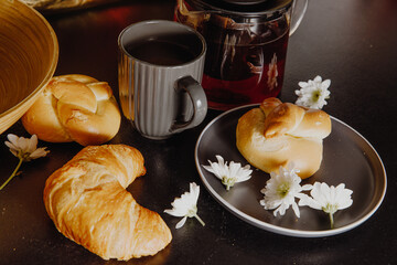 bakery products arranged on a black table with a cup of tea. croissant with sweet baking on the black table and with small white flowers. sweet pastries on a gray background with a gray plate 