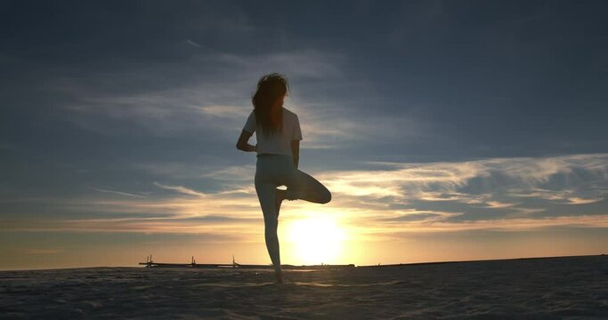 On the general plan of the beach at sunset, the yogi stands with her back to the camera and makes a tree pose, resting the foot of one foot on the knee of the other, then she wraps her legs together