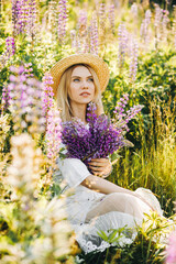 Serious lady in a hat sitting in the middle of a lupine field holding a bouquet of flowers in her hand
