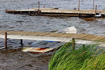 Fototapeta premium Abandoned old drowned boat in the water under a wooden pier on windy grass background, water landscape after storm