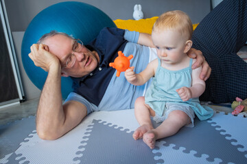 Adorable red-haired newborn sitting on floor and playing toy. Happy grandfather in eyeglasses and...