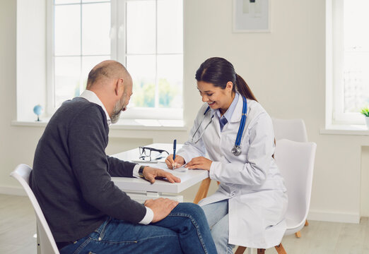 Female Doctor Writing Medication Order For Senior Patient In Clinic. Elderly Man On Visit To His Healthcare Provider