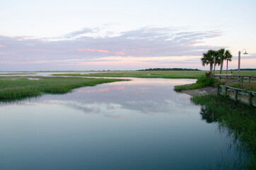 A peaceful, tranquil scene of a mirror lake with reeds, grass, trees, a street lamp, and a distant hill. The sky and it's reflection have a pink sunset hue.